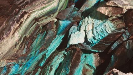 Close-up of jagged rock layers with shimmering turquoise, teal, and brown mineral streaks creating a textured, iridescent surface.